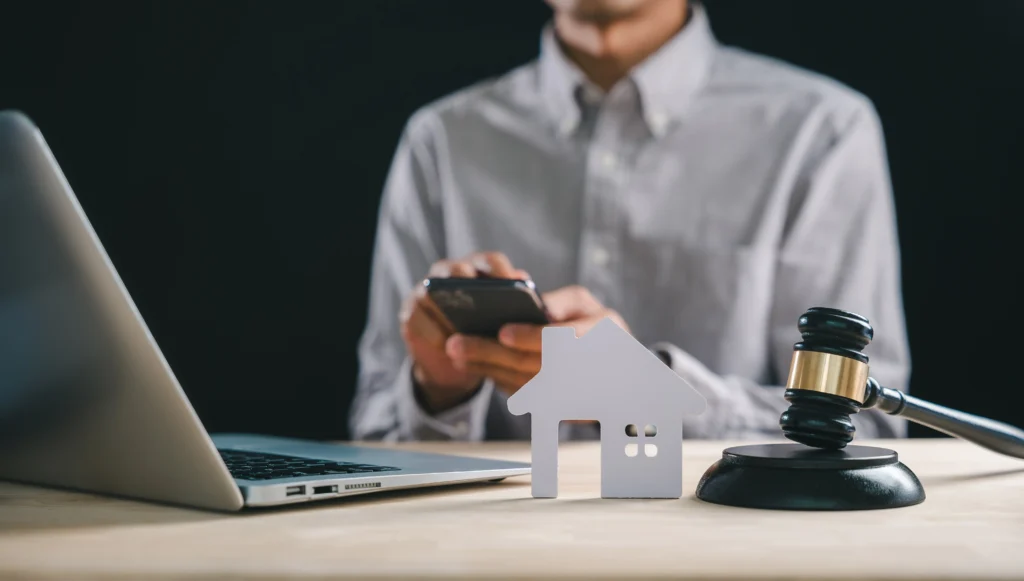 Gavel and house silhouette beside a laptop and person using a phone